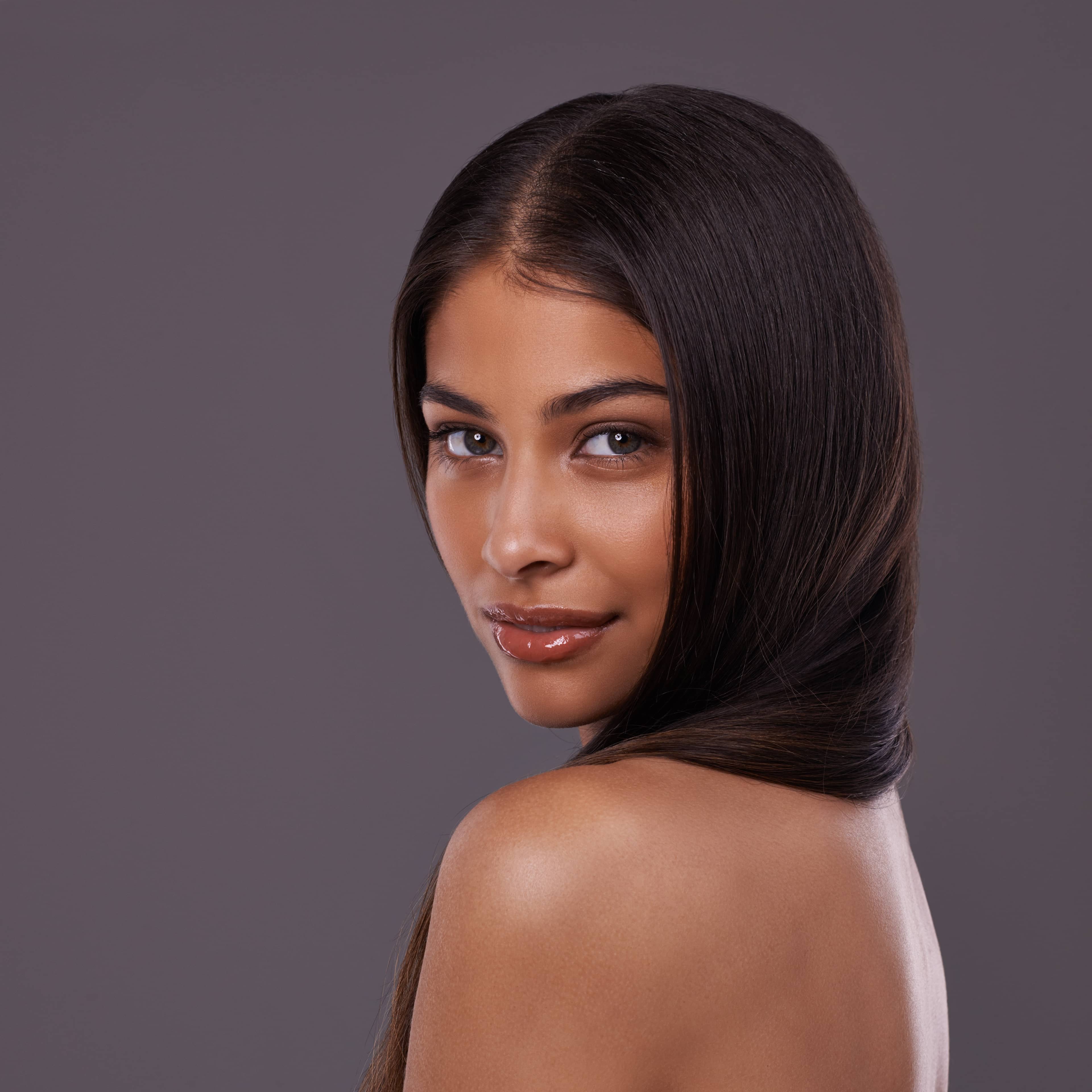 Portrét mladé ženy s dlouhými, rovnými tmavými vlasy a lesklými rty, která se jemně usmívá a dívá přes rameno do kamery na tmavě šedém pozadí.
