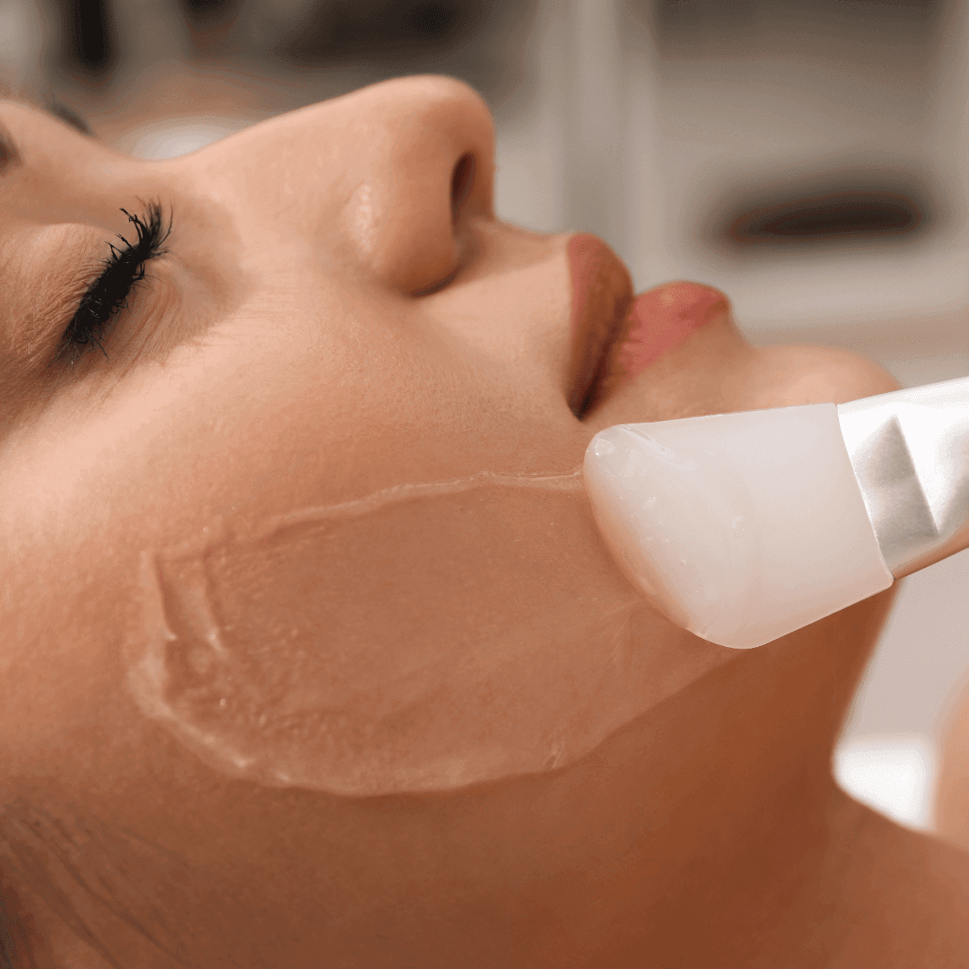 Žena leží se zavřenýma očima, zatímco jí kosmetička nanáší štětcem průhlednou masku na tvář během kosmetického ošetření.