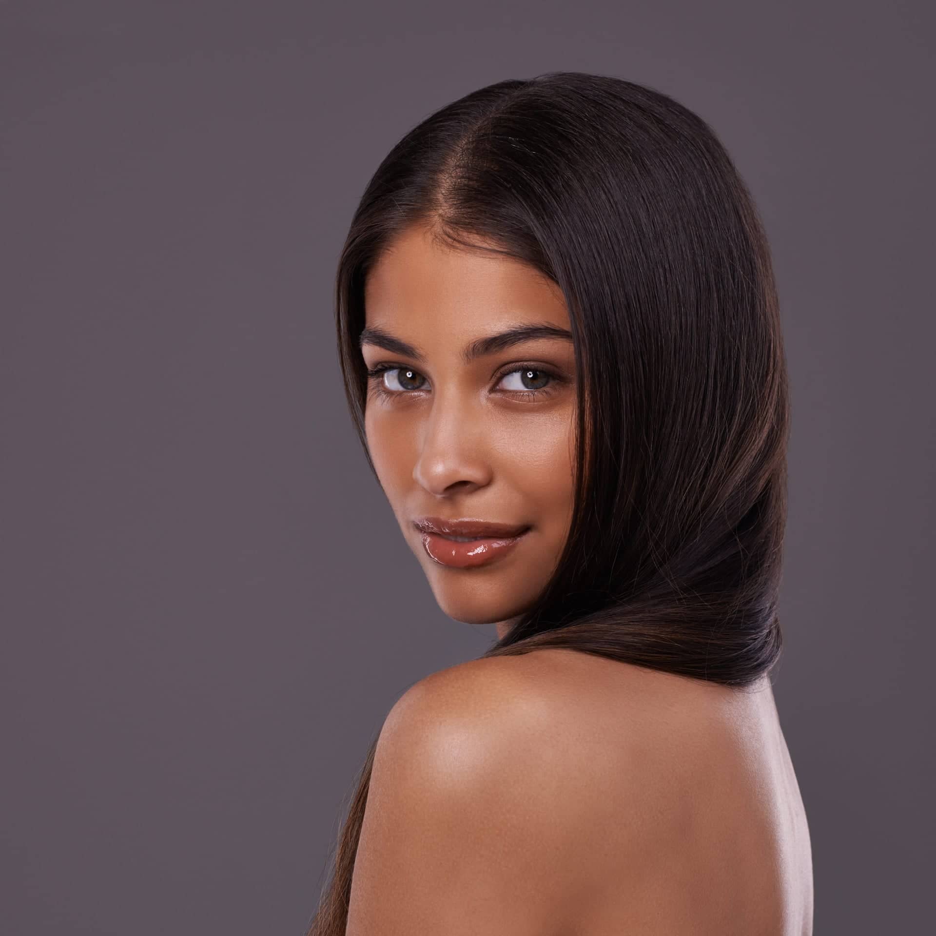 Portrait of a young woman with long, straight dark hair and glossy lips, smiling softly and looking over her shoulder at the camera against a dark gray background.