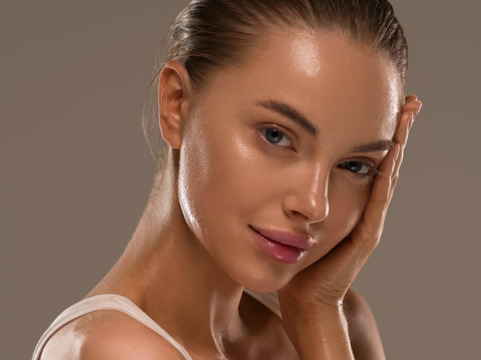 Portrait of a young woman with shiny, healthy skin and tied‑back hair, with soft natural makeup, gently touching her face and looking into the camera against a neutral brown‑gray background.