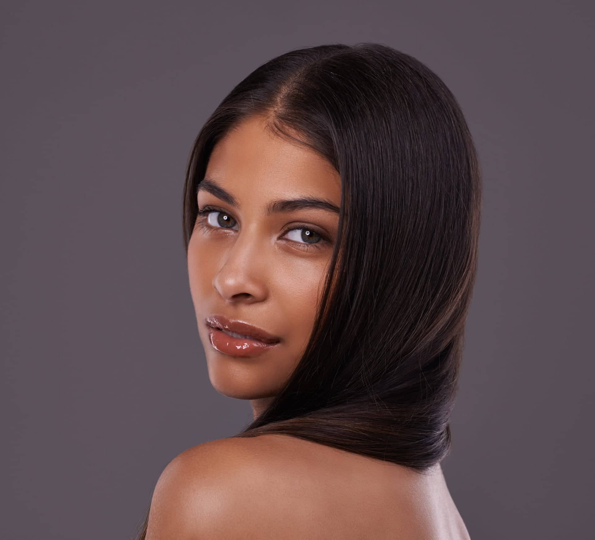 Portrait of a young woman with smooth, shiny dark hair and natural makeup, gently looking over her shoulder at the camera against a dark gray background.