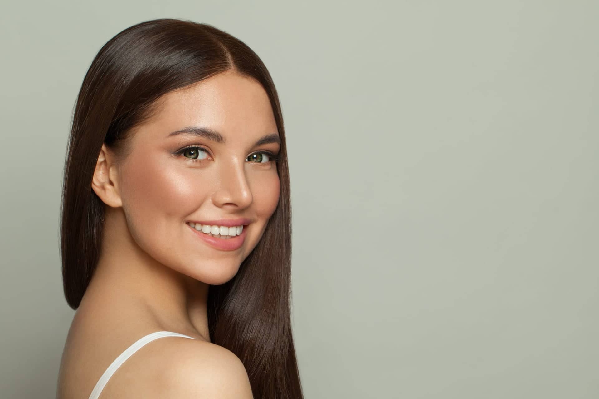 Portrait of a young woman with long brown hair and soft makeup, smiling and looking into the camera against a neutral gray‑green background.
