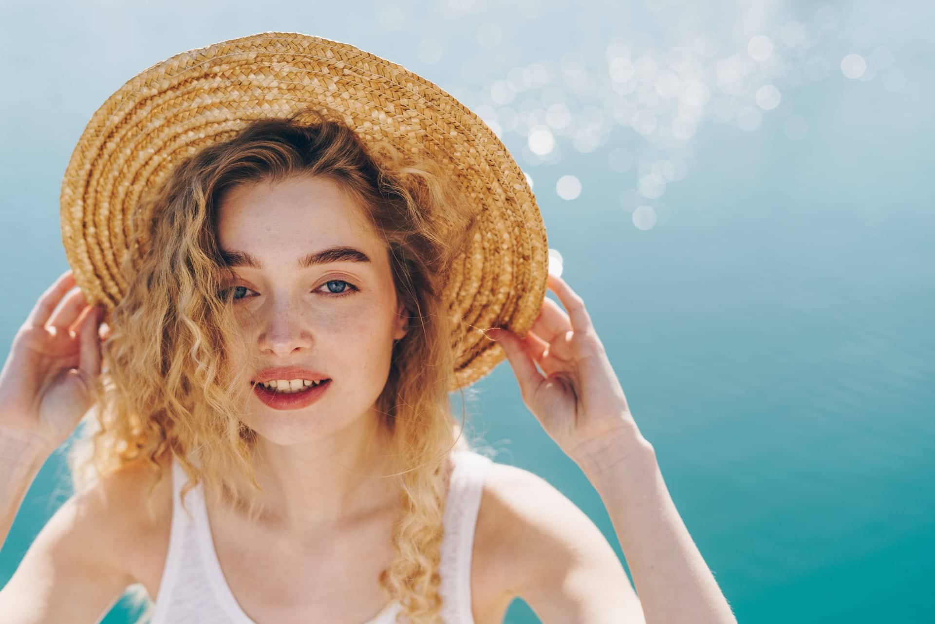 A young woman with curly blonde hair and a straw hat smiles into the camera by the water, wearing a white top, with sunlight sparkling on the sea surface in the background.