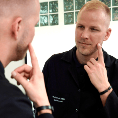 Dermatologist MUDr. Martin Bortlík from Skinity Clinic looking into a mirror and pointing to his face.