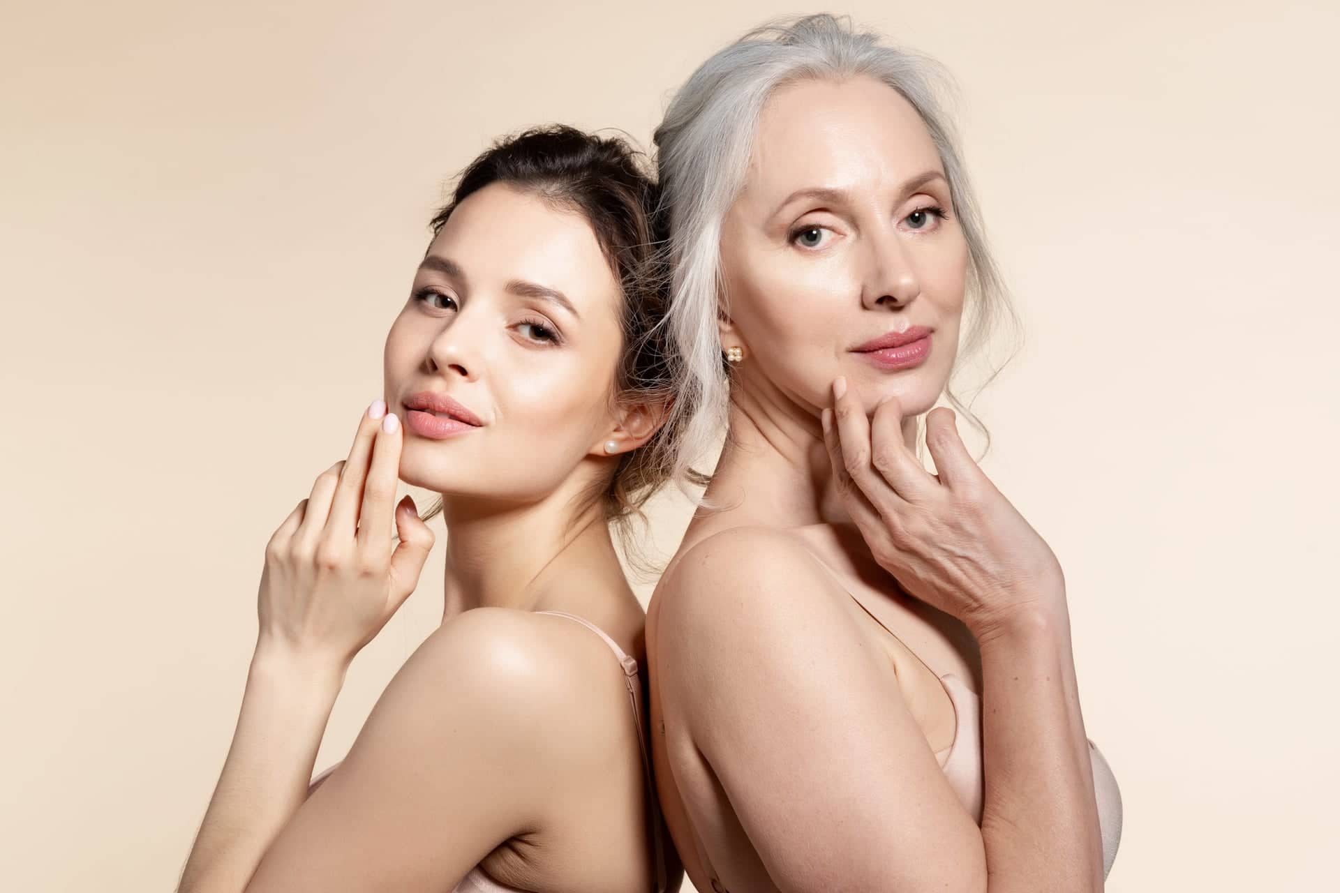 Portrait of two women standing back-to-back against a neutral beige background – a younger woman with dark hair and an older woman with gray hair, both with gentle smiles and natural makeup, touching their faces and exuding elegance and confidence.