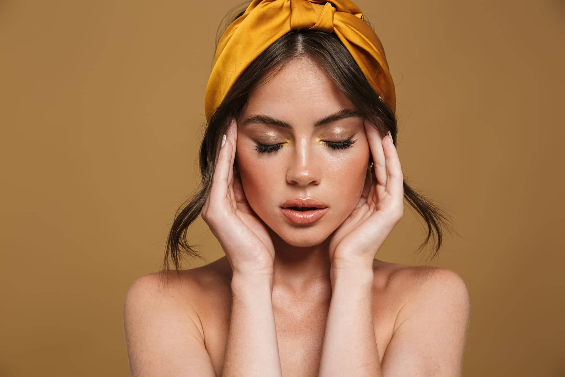 Portrait of a young woman with soft makeup and closed eyes, gently holding her face in her hands. She has brown hair in a bun and a bright yellow headband matching the warm beige‑brown background.