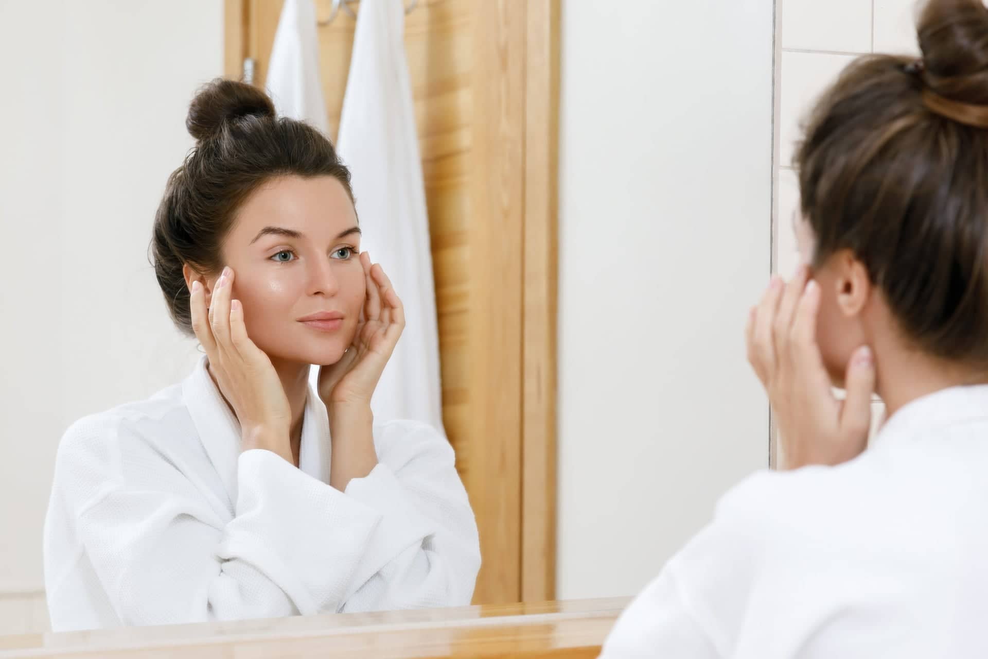 A young woman in a white robe stands in front of a mirror in the bathroom and gently touches her face as she examines her skin. She has dark hair in a bun and a calm, content expression.