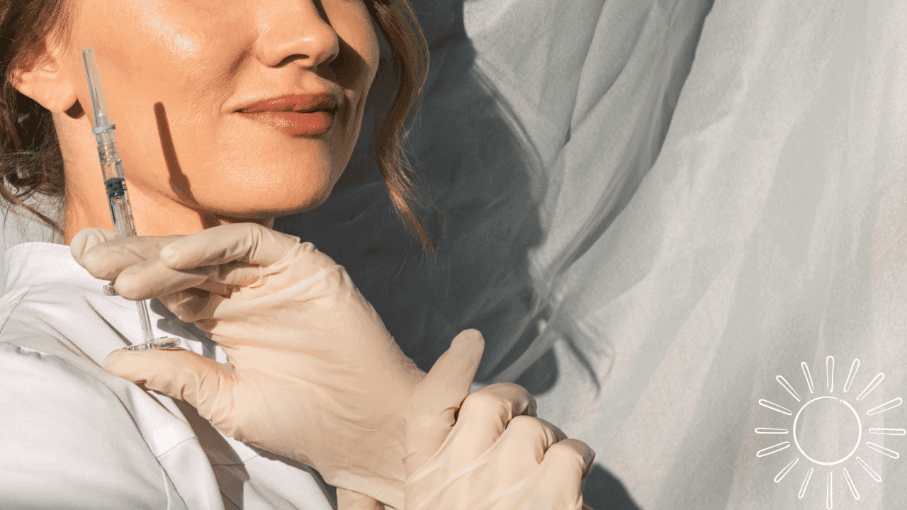 Close-up of a woman in a white coat wearing latex gloves and holding a syringe. Her expression is professional and calm, while her face and the bright white background are illuminated by sunlight.