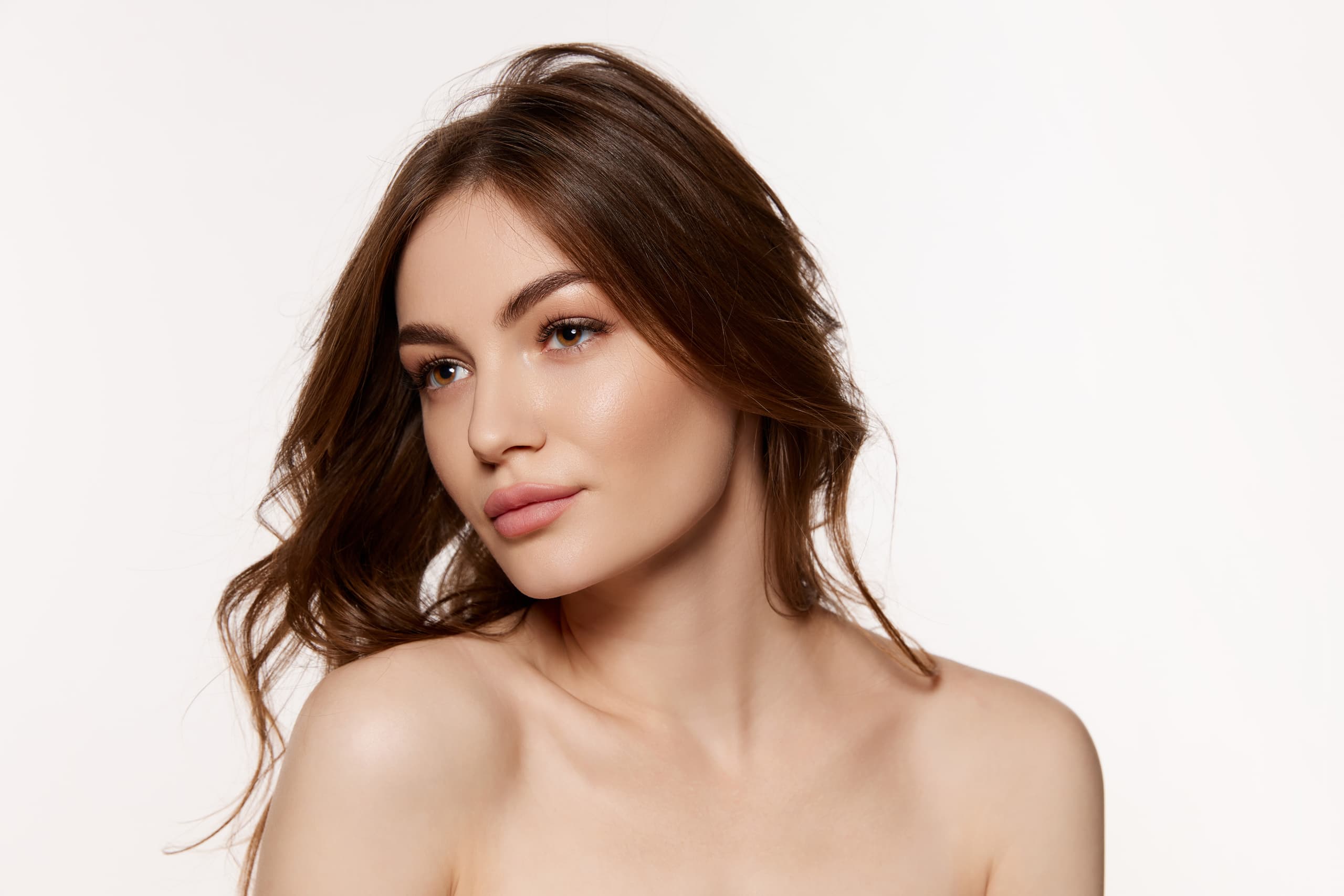 Portrait of a young woman with soft makeup and wavy brown hair looking to the side. Her skin is smooth and radiant, and she poses against a beige background.