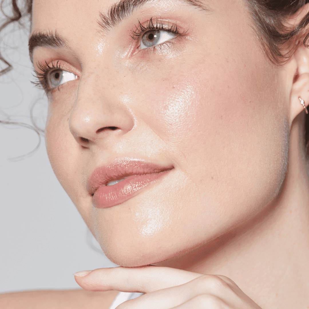 Close-up portrait of a woman with soft makeup and healthy, glowing skin. She looks into the distance with a gentle smile, supporting her chin with her hand.