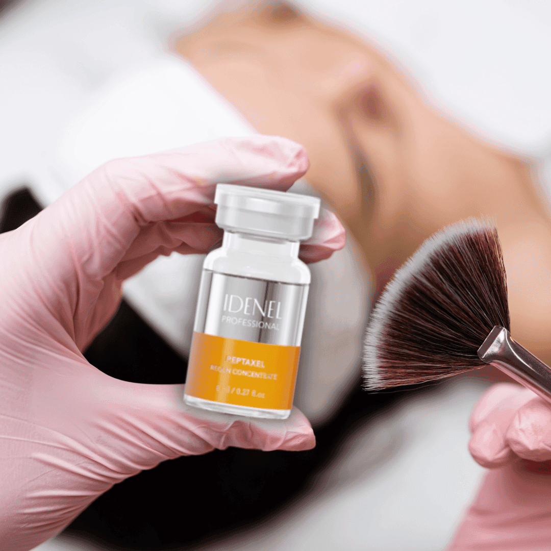Kosmetička v růžových rukavicích drží lahvičku s přípravkem IDENEL Professional Peptaxel Regen Concentrate a vějířovitý štětec, připravená k aplikaci na pleť klientky ležící na pozadí.